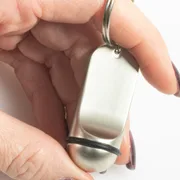 Hand, die einen mattsilbernen, länglichen Schlüsselanhänger mit schwarzem Gummiring an einem Schlüsselring hält, vor weißem Hintergrund.